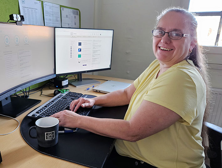En leende Annmarie Fogelberg sitter vid ett skrivbord som har två bildskärmar och ett tangentbord.